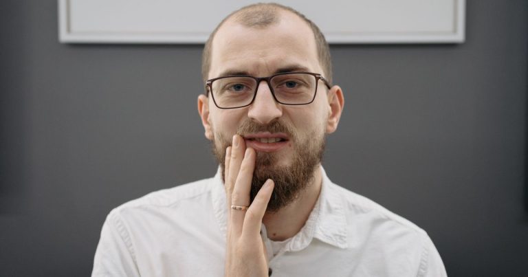 Man with glasses and a beard pressing his fingers against his cheek in discomfort
